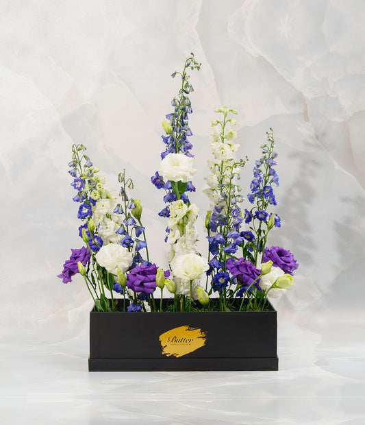 A beautifully composed floral display in a rectangular dark box, featuring tall featuring exotic delphiniums and lisianthus. Together, they evoke the feel of a wild alpine field.