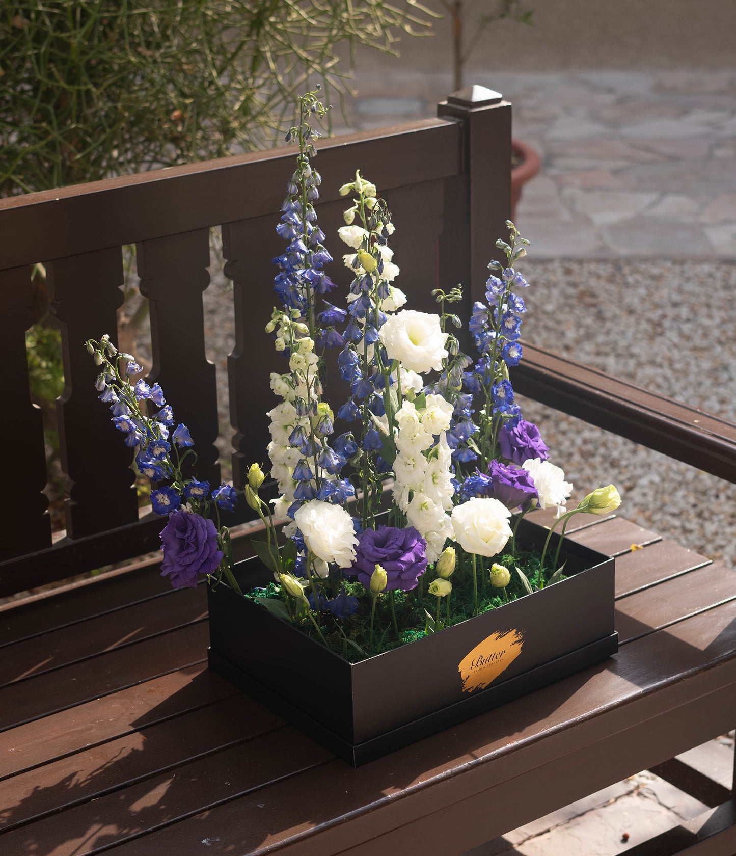 A close-up of the front of the arrangement, showcasing the delicate textures and contrasting colors of the purple delphiniums and white lisianthus against the dark container.