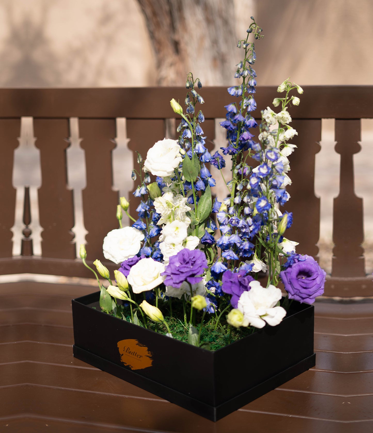 Butter Daphne’s Field bouquet with purple delphiniums and white lisianthus in a stylish dark box—nostalgic, natural, and refreshing.