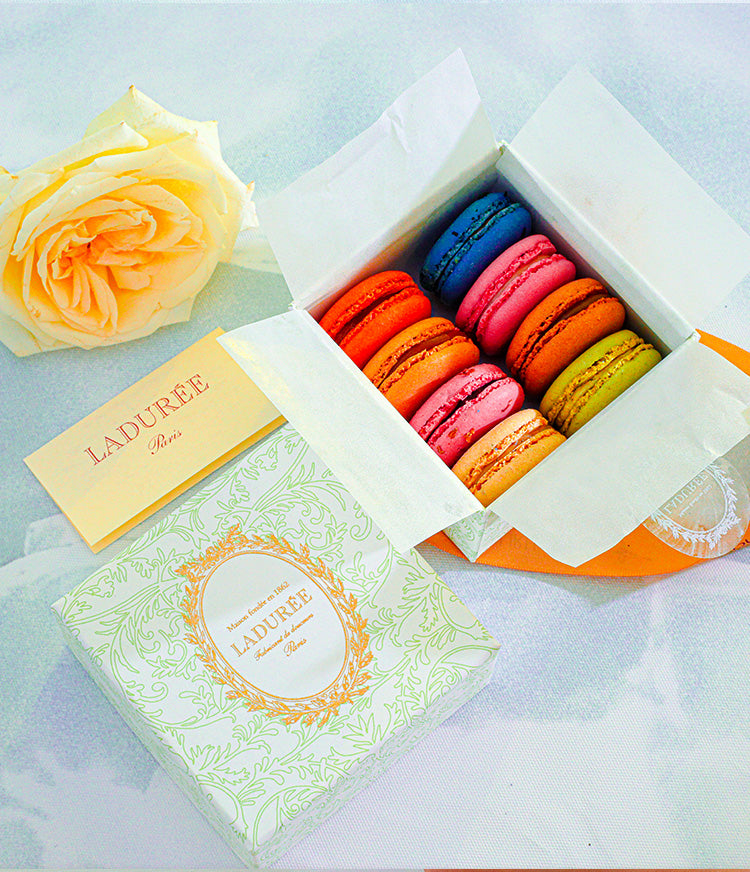 A pastel green Ladurée Arabesque Box featuring an elegant gold embossed design, filled with colorful assorted macarons arranged neatly inside.