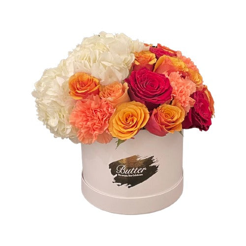 A round matte white box in medium size, filled with a bright and cheerful mix of hydrangeas, roses, baby roses, and carnations in shades of red, orange, and yellow.