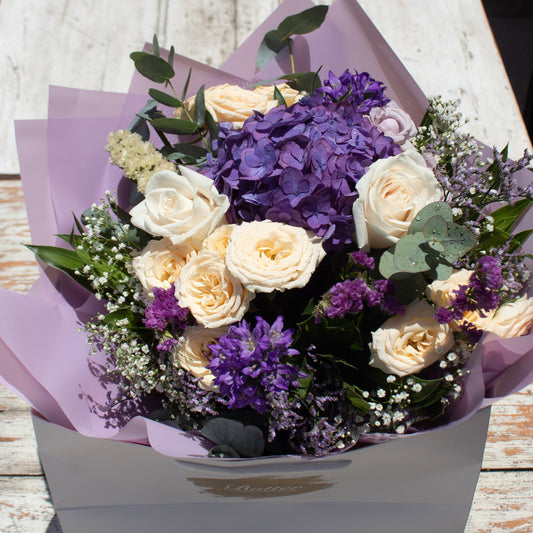 A luxurious bouquet featuring a lush cluster of deep purple hydrangeas surrounded by cream-colored roses, delicate purple statice, baby’s breath, and greenery, elegantly wrapped in pale lavender paper inside a branded box.