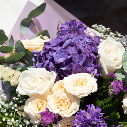 Close-up of a sophisticated floral arrangement: vibrant purple hydrangeas at the center, cream roses, purple filler flowers, and eucalyptus sprigs nestled in lavender-toned wrapping.