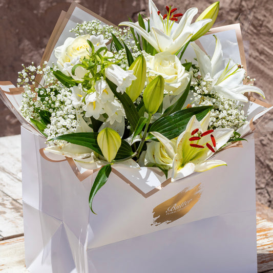 A fresh flower bouquet featuring white blooms, green leaves, and red accents, delivered in a matte white bag.