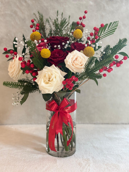 A festive floral arrangement featuring red and white roses, golden craspedia, vibrant berries, and rich greenery, all elegantly presented in a clear vase with a ribbon tied to it, that says the word “BUTTER”. This bouquet captures the magic of the season, bringing warmth and elegance to any space.