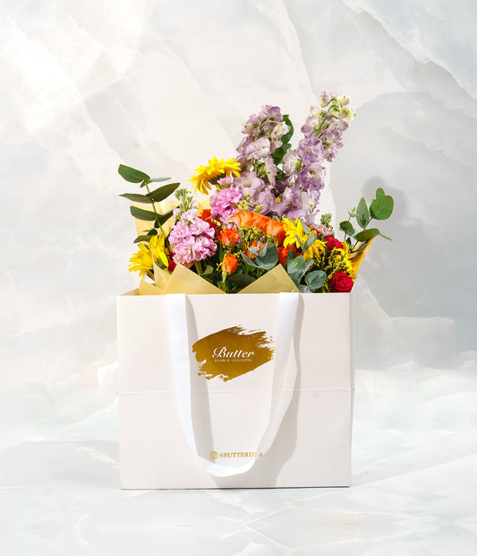A white paper bag with a Butter brand tag, filled with a vibrant arrangement of multicolored flowers including sunflowers and purple flowers.