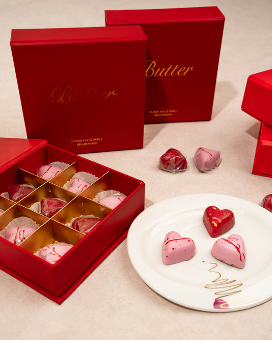 A vibrant red gift box with BUTTER branding is displayed in the background. In the foreground, a selection of heart shaped chocolates with delicate butterfly-shaped decorations is arranged on a plate.