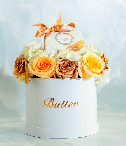 A white hat-box with 24K Gold dusted roses, accompanied by a box of Ladurée macarons, presented with a bow and the brand 'Butter' visible on the box.