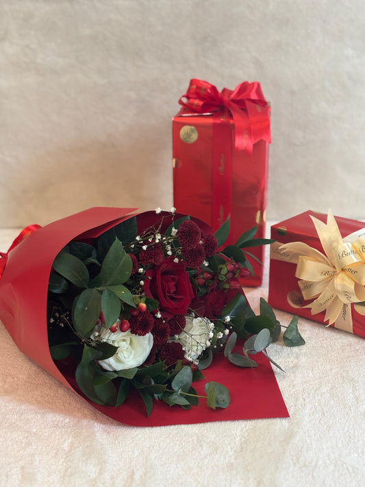 A vibrant red Christmas crimson bouquet of flowers including red roses, white spray roses, and greenery, presented alongside a red gift box with a gold ribbon.