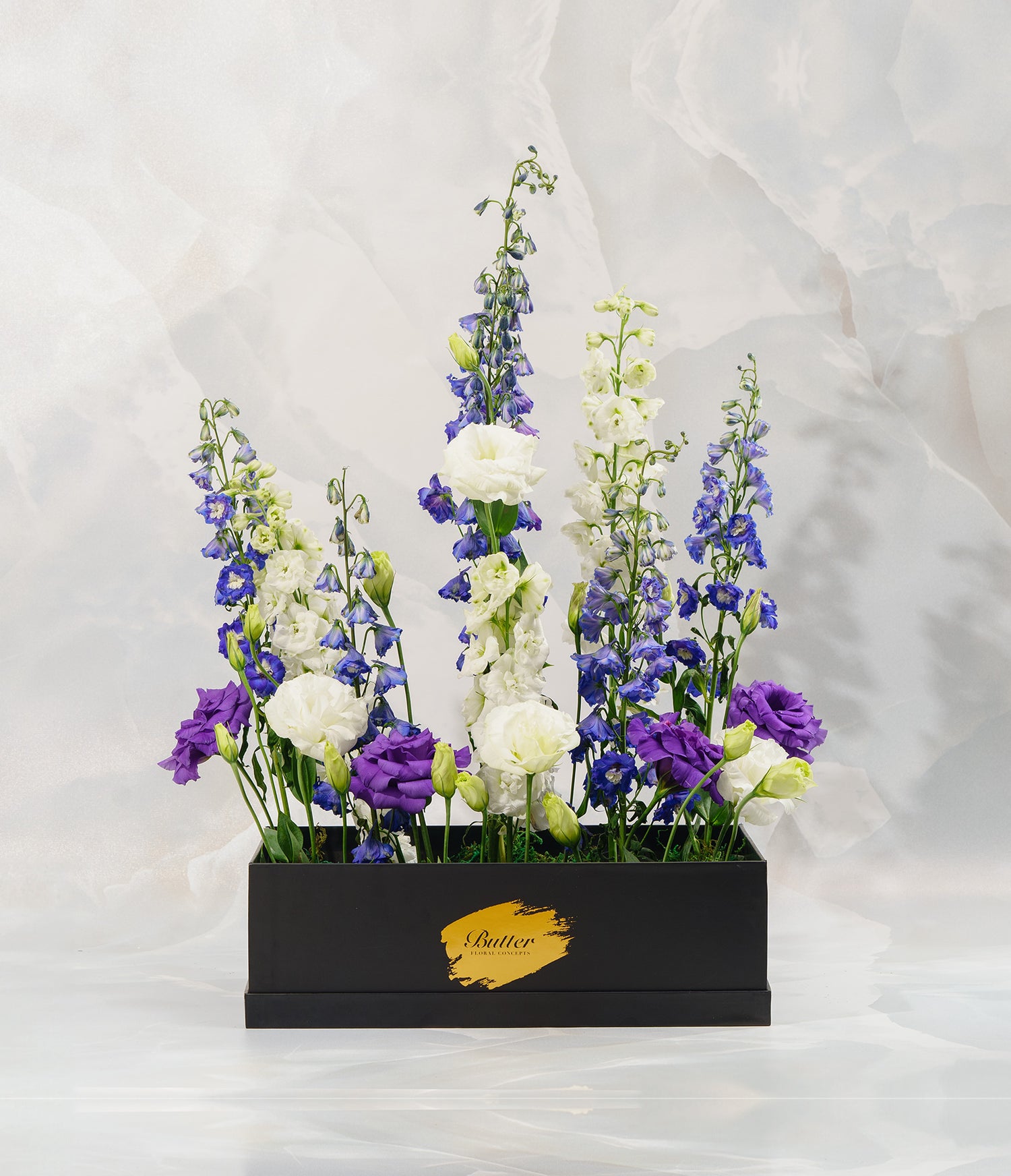 A beautifully composed floral display in a rectangular dark box, featuring tall featuring exotic delphiniums and lisianthus. Together, they evoke the feel of a wild alpine field.