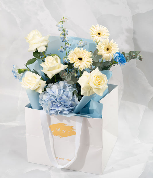 A floral arrangement featuring a mix of fresh seasonal flowers flowers including roses and others in shades of white, blue, and pearl, presented in a white paper bag with a blue ribbon.