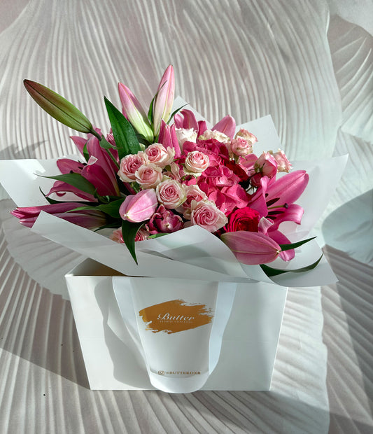 A vibrant pink floral bouquet containing pink roses, lilies, and hydrangeas, wrapped in a white paper with a gold logo.