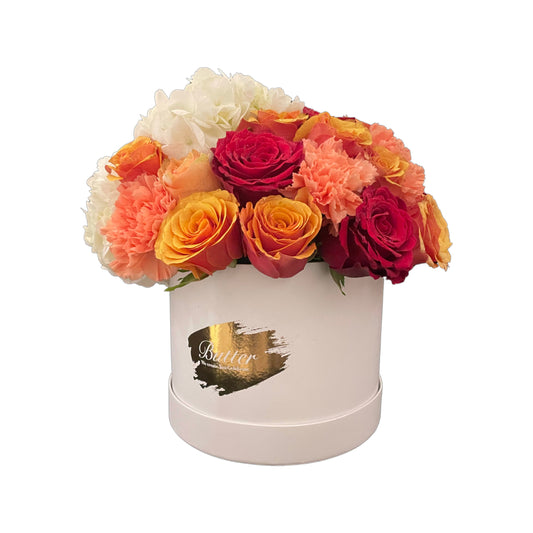 A medium round matte white box filled with an arrangement of hydrangeas, roses, baby roses, and carnations in shades of orange, red, and yellow.