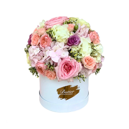 A dome-shaped floral arrangement with pastel pink hydrangeas, baby peach, lilac, and pearl white roses in a matte white box with the brand name 'Belle' visible.