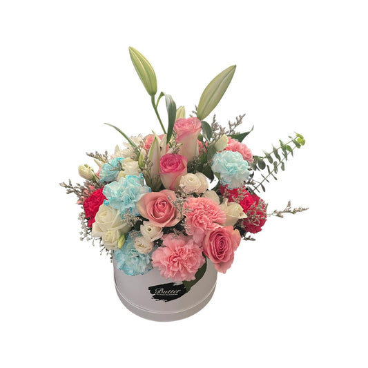 A round matte white box containing an arrangement of fresh flowers including roses, baby roses, carnations, lilies, and lavender in a variety of pastel colors.