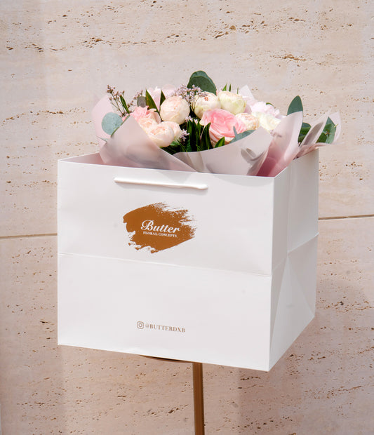 A bouquet of soft pink flowers consisting of baby peonies, hydrangeas, and garden roses, arranged in a white paper bag with a logo on the front.