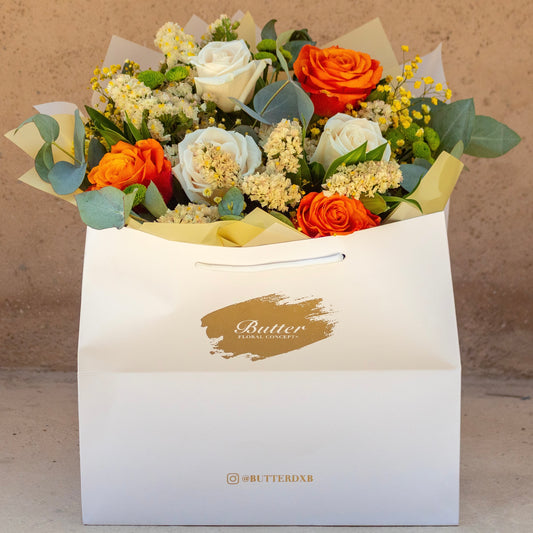 A bouquet of fresh flowers featuring orange roses, arranged in a white bag with the brand name 'Butter' printed on it.