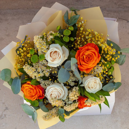A bright and cheerful flower arrangement striking orange roses mixed with softer, white-toned blooms and touches of greenery. The bouquet is elegantly arranged in a Butter-branded matte white bag, creating a vibrant yet refined look.